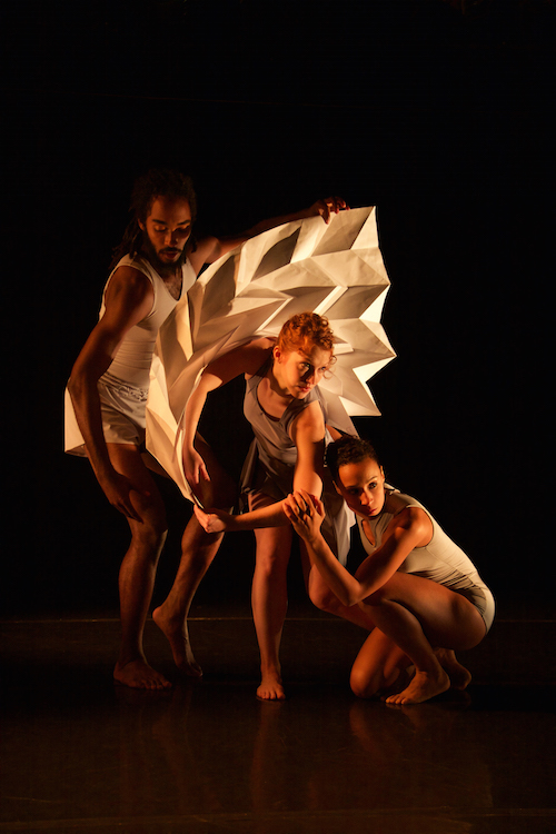 The trio of dancers huddle around a square-shaped accordion looking paper